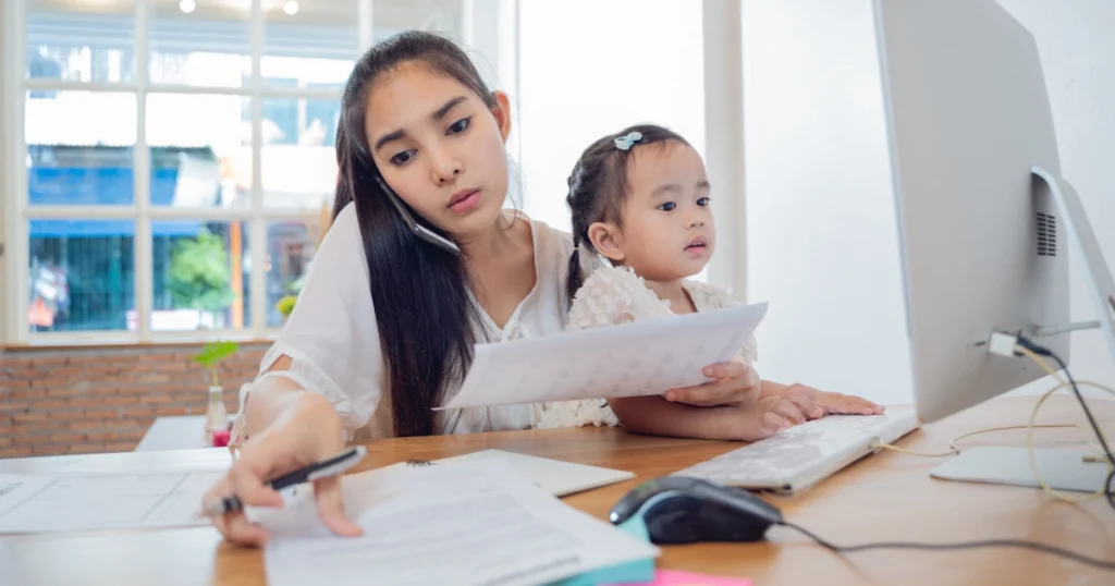 Parent on phone reviewing homeschool paperwork with child at home desk