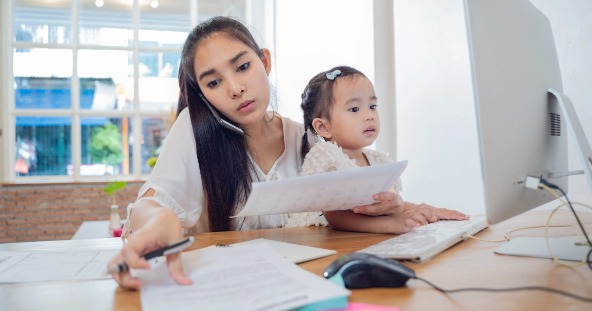 Parent on phone reviewing homeschool paperwork with child at home desk
