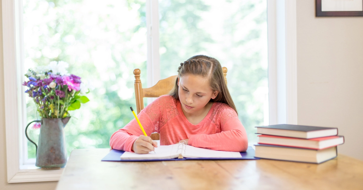 Upper elementary student writing in a binder at a table with stacked books during homeschool study time