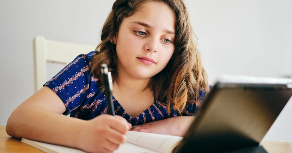 Child writing in a notebook while using a tablet for a homeschool lesson, showing a balanced online and offline learning approach