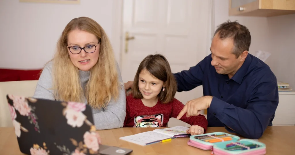 Parents helping their daughter with homeschool lessons at the kitchen table while working together in a realistic home learning environment.
