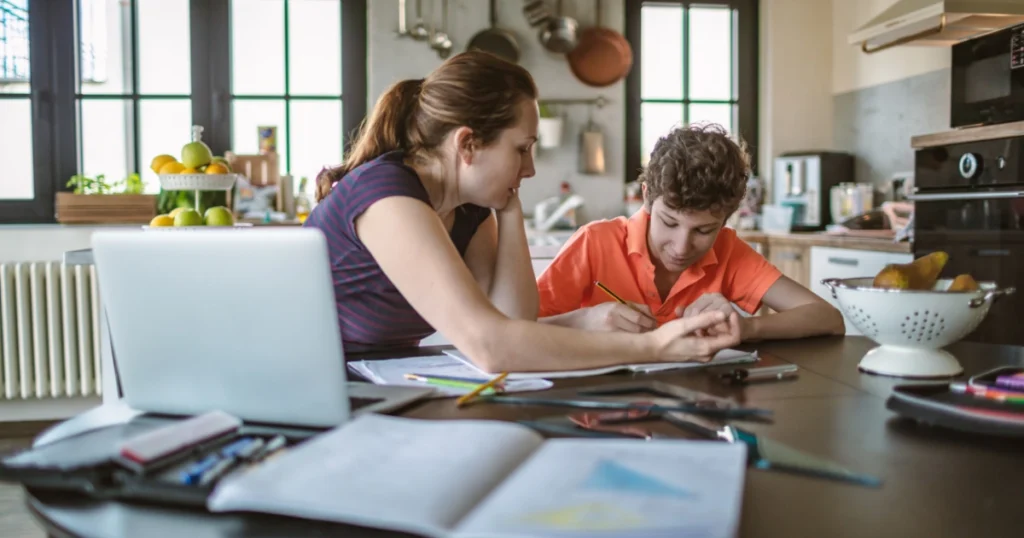 Parent helping a child with homeschooling at a busy kitchen table surrounded by books, papers, and school materials.