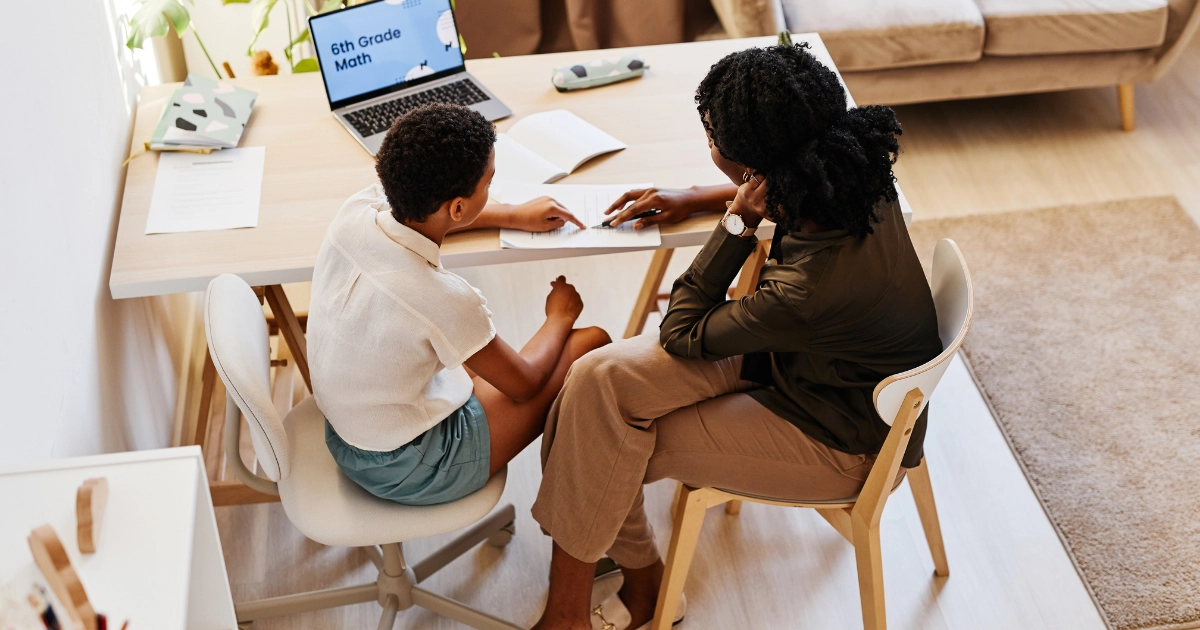 Parent homeschooling a child at a kitchen table, working together on a math lesson in a home learning environment.