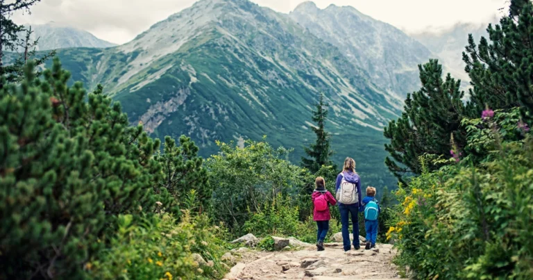 Parent and two children hiking along a mountain trail surrounded by trees with towering peaks in the distance.